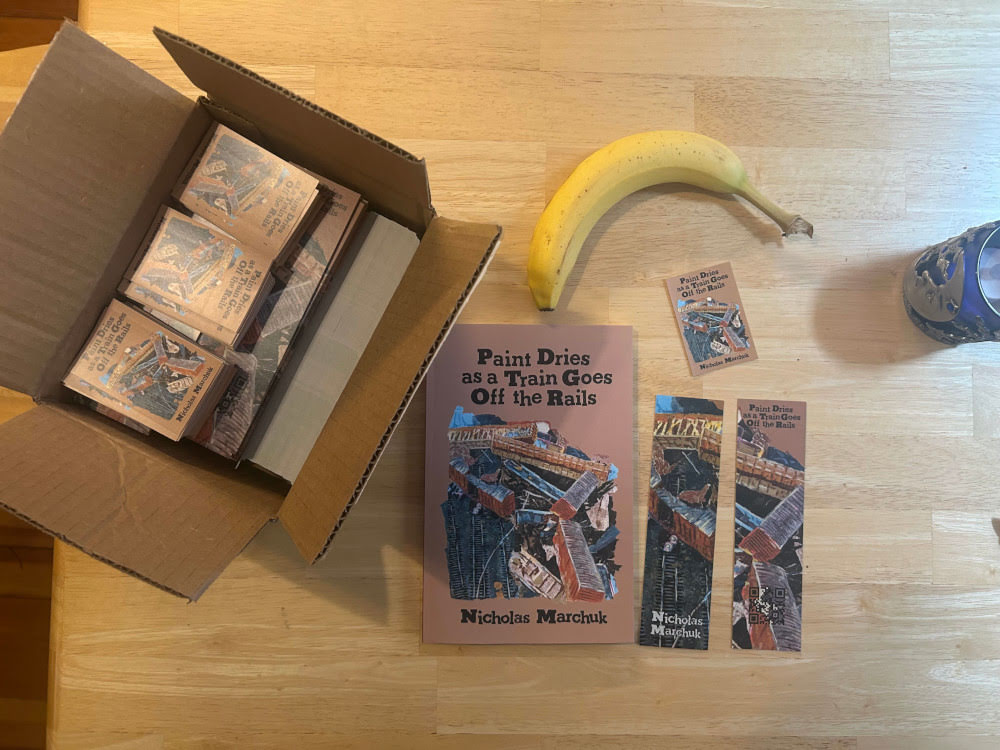 A copy of Paint Dries as a Train Goes Off The Rails on a table. The cover depicts a train derailment and pileup in a mixed media collage. There are bookmarks, stickers, as well as a single banana